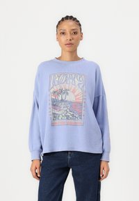 Lichtblauwe oversized sweatshirt met een grafische print die een strandscène, palmbomen en het woord "ROXY" in gestileerde tekst toont.