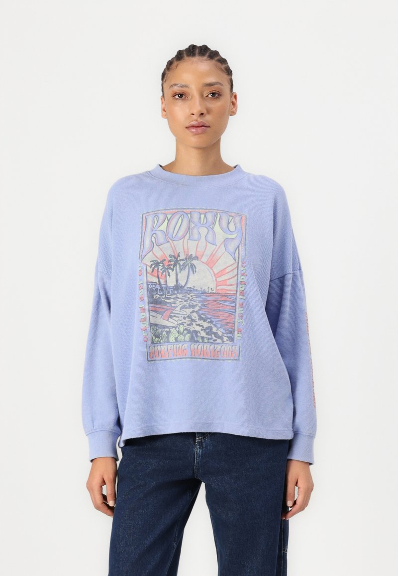Lichtblauwe oversized sweatshirt met een grafische print die een strandscène, palmbomen en het woord "ROXY" in gestileerde tekst toont.