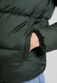 Handen i sidofickan på en mörkgrön vadderad pufferjacka med elastiska ärmslut, bärs över ljusblå denimjeans.