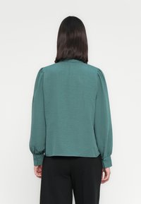 Τιρκουάζ μακρυμάνικη μπλούζα με υφή, συγκεντρωμένους ώμους και κουμπωτές μανσέτες, συνδυασμένη με μαύρο παντελόνι.