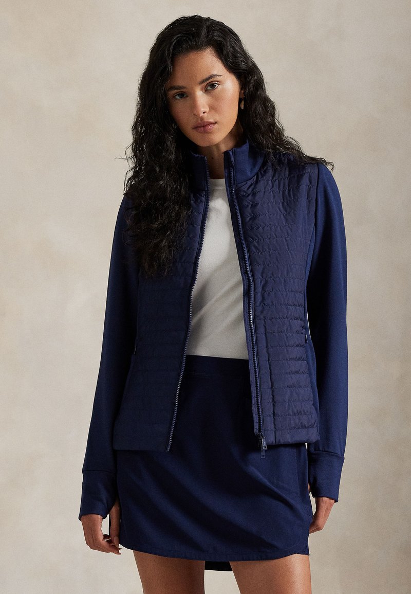 Μπλε ναυτικό μπουφάν με καπιτονέ μπροστινό μέρος και λεία μανίκια, με ψηλό γιακά και κλείσιμο με φερμουάρ. Συνδυασμένο με μια ταιριαστή μπλε ναυτική φούστα.