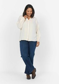 Camicia crema con pieghe verticali, chiusura a bottoni frontale e maniche lunghe, abbinata a jeans blu scuro e stivaletti marroni.