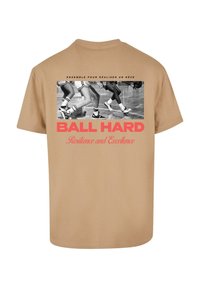 Beige bomulds T-shirt med et sort-hvidt grafik af basketballspillere og rød tekst "BALL HARD" med "Modstandsdygtighed og Ekscellence" nedenunder.