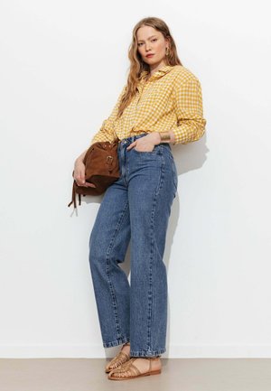 Femme portant une chemise jaune à carreaux, jean bleu taille haute, sandales tressées marron, et tenant un sac en suède marron sur fond blanc.