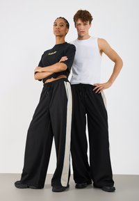 Melni platajā stila bikses ar krēmkrāsas sānu svītru un zilu akcentu, valkātas ar melnu T-kreklu un baltu topu, abi ir ikdienišķi stili.