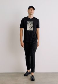 Tricou negru cu mânecă scurtă, cu un print grafic care reprezintă un câine, asociat cu pantaloni negri și mocasini, purtat de un model masculin în picioare.