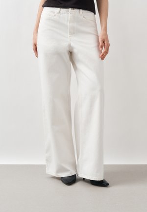 Persoană purtând blugi albi cu talie înaltă și croială largă pe picior și pantofi negri cu vârf ascuțit, stând pe un fundal gri simplu.