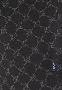 Černá látka s texturovaným vzorem propletených kruhů a květinových akcentů, s malou modrou štítkem s nápisem "JOOP!" na okraji.