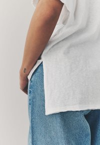 White cotton t-shirt features a relaxed fit and side slits, paired with light blue denim jeans in a faded wash with subtle texture.