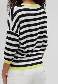 Gestreifter schwarz-weißer Pullover mit einem limettengrünen Kragen, drei Viertelärmeln und lockerer Passform. Dekorative Akzente an der Hose.