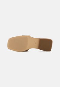 Chaussure à talons hauts beige avec une semelle en caoutchouc texturée. Le talon est large et en forme de bloc, avec des lanières en cuir lisse sur le dessus.