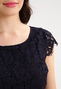 Haut en dentelle bleu marine avec des motifs floraux, col rond et délicates manches courtes à bords festonnés. Texture douce et design complexe.