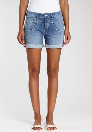 Vrouw met opgerolde blauwe spijkershorts en witte slippers, staande tegen een effen witte achtergrond.