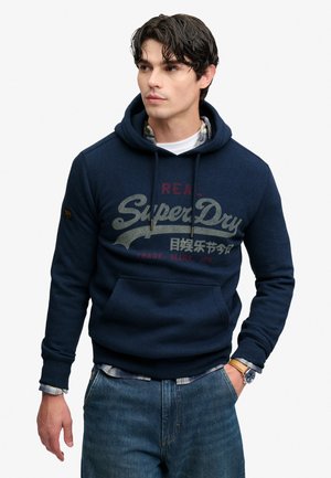 Młody mężczyzna w granatowej bluzie z kapturem SuperDry na koszuli w kratę, niebieskich dżinsach i zegarku na rękę, patrząc na bok z jedną ręką w kieszeni.