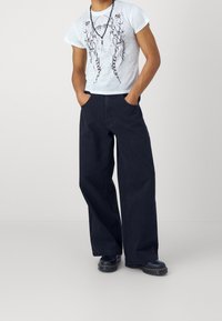 Bílý grafický tričko s černým abstraktním potiskem, spárováno s širokými tmavými kalhotami a černou obuví na šněrování. Neformální outfit, uvolněný střih.