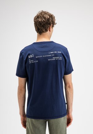 Man met krullend haar die een marineblauw T-shirt draagt met witte opdruk op de bovenrug, staande tegen een egale achtergrond.