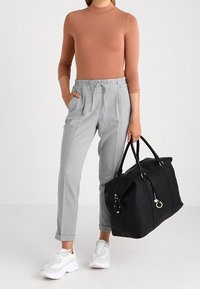 Sort duffel bag med lynlås og metalliske detaljer. Modellen bærer en tætsiddende brun bluse, grå bukser og hvide sneakers.