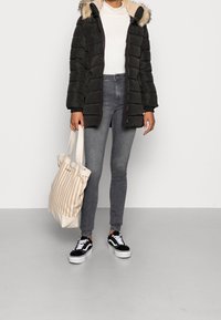 Černá péřová bunda s kapucí obšitou kožešinou, bílý žebrovaný top, šedé skinny džíny a černo-bílé tenisky, držící bé�žovou pruhovanou tašku.