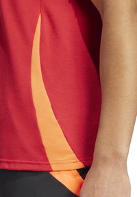 Rode sportshirt met een gestructureerde stof en een diagonale oranje accent aan de linkerkant. Zwarte shorts met oranje rand zichtbaar.