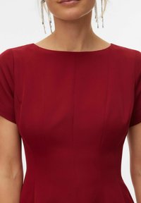 Crvena haljina s izrezom u obliku čamca i kratkim rukavima. Izrađena od glatkog materijala, s diskretnim vertikalnim šavovima. Naušnice s padajućim lancima.