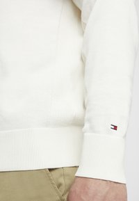 Hellbeiger Strickpullover mit geripptem Kragen und Bündchen. Verfügt über ein kleines Logo am Ärmel. Weiche Textur, klassischer Schnitt und minimalistisches Design.