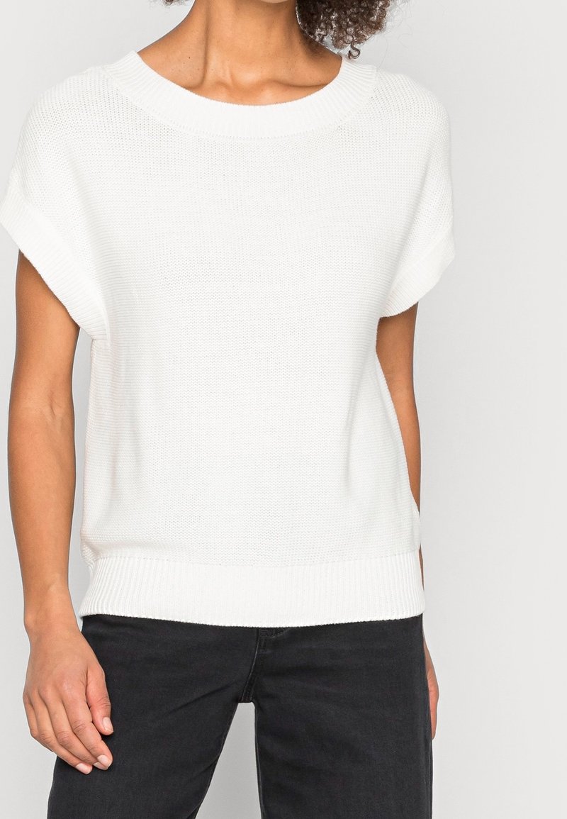 Femme portant un haut en maille blanc à manches courtes avec ourlet et encolure côtelés, associé à un pantalon noir sur un fond clair.