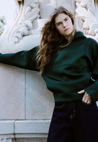 Tmavě zelená mikina s kapucí v uvolněném střihu, vyrobená z texturované látky a bezešvého provedení. Kombinována s tmavými džínami s vysokým pasem.