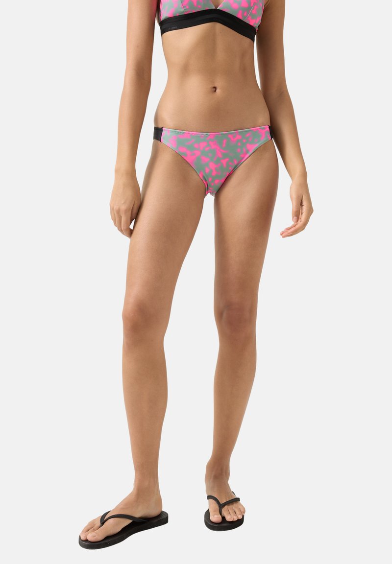 Frau trägt pink-grau gemusterte Bikinihose und schwarze Flip-Flops, stehend vor einem schlichten hellen Hintergrund.