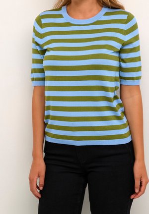Vrouw draagt een nauwsluitende trui met korte mouwen met horizontale blauwgroene strepen, gecombineerd met een zwarte broek, staand tegen een effen muur.