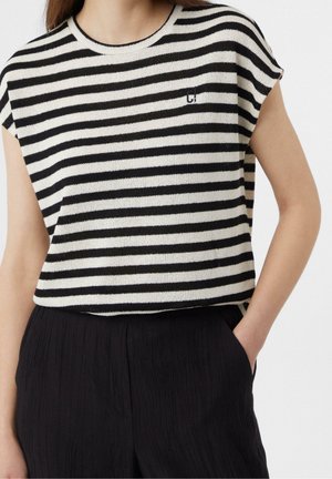 Frau trägt ein schwarz-weiß horizontal gestreiftes ärmelloses Oberteil mit schwarzen Hosen, eine Hand in der Tasche, vor weißem Hintergrund.