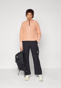Giacca zip-up rosa chiaro pelosa, pantaloni cargo neri con tasche e uno zaino nero. L'outfit è completato da scarpe beige chiare.
