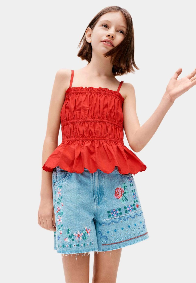 Niña joven con una blusa sin mangas roja fruncida y shorts de mezclilla azul claro con bordado floral colorido sobre un fondo blanco.