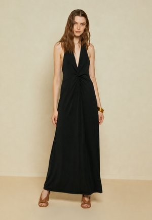 Donna dai capelli castani ondulati che indossa un abito lungo nero senza maniche con collo ad halter e tacchi aperti marroni, in piedi davanti a uno sfondo beige semplice.