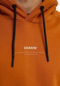 Oranje hoodie met zwarte trekkoorden, een gestructureerd oppervlak en bedrukte tekst met de merknaam "CHASIN'" en een beschrijvende zin eronder.