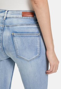 Jean en denim bleu clair avec une texture lisse, comportant une étiquette en cuir portant l'inscription "BETTY BARCLAY" et deux poches arrière.