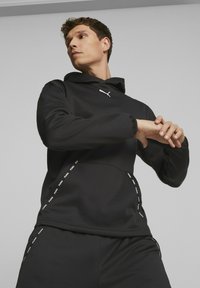 Sudadera negra con tejido texturizado, logo en blanco, puños elásticos y panel lateral con el nombre de la marca repetido. La pose del modelo presenta las manos al frente.