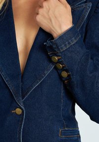 Blazer in denim blu scuro con profondo scollo a V, spalle strutturate e dettagli in bottone di ottone sui polsini. La cucitura a vista aggiunge particolari.