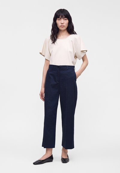 Femme aux cheveux noirs et raides portant un chemisier beige à manches courtes, un pantalon court taille haute bleu marine et des chaussures plates noires, debout, une main dans la poche.
