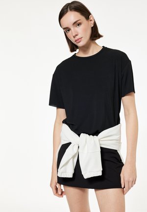 Jonge vrouw met kort bruin haar die een losse zwarte T-shirtjurk draagt en een wit sweatshirt om haar taille heeft gebonden.