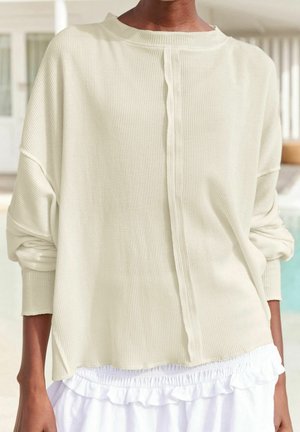 Personne portant un haut ample en tissu gaufré à manches longues de couleur blanc cassé avec les manches retroussées et une jupe ou un short blanc à volants, dans un endroit en intérieur.