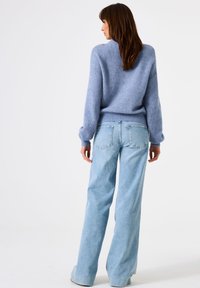 Frau mit braunen Haaren, die einen hellblauen, flauschigen Pullover und lockere, hellblaue Jeans trägt, steht mit dem Gesicht zur linken Seite vor einem weißen Hintergrund.