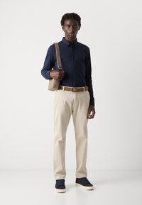 Marineblauwe lange mouwen shirt, beige broek, bruin riem en marineblauwe sneakers. Model houdt een lichtbruine tas vast; gladde texturen, op maat gemaakte pasvorm.