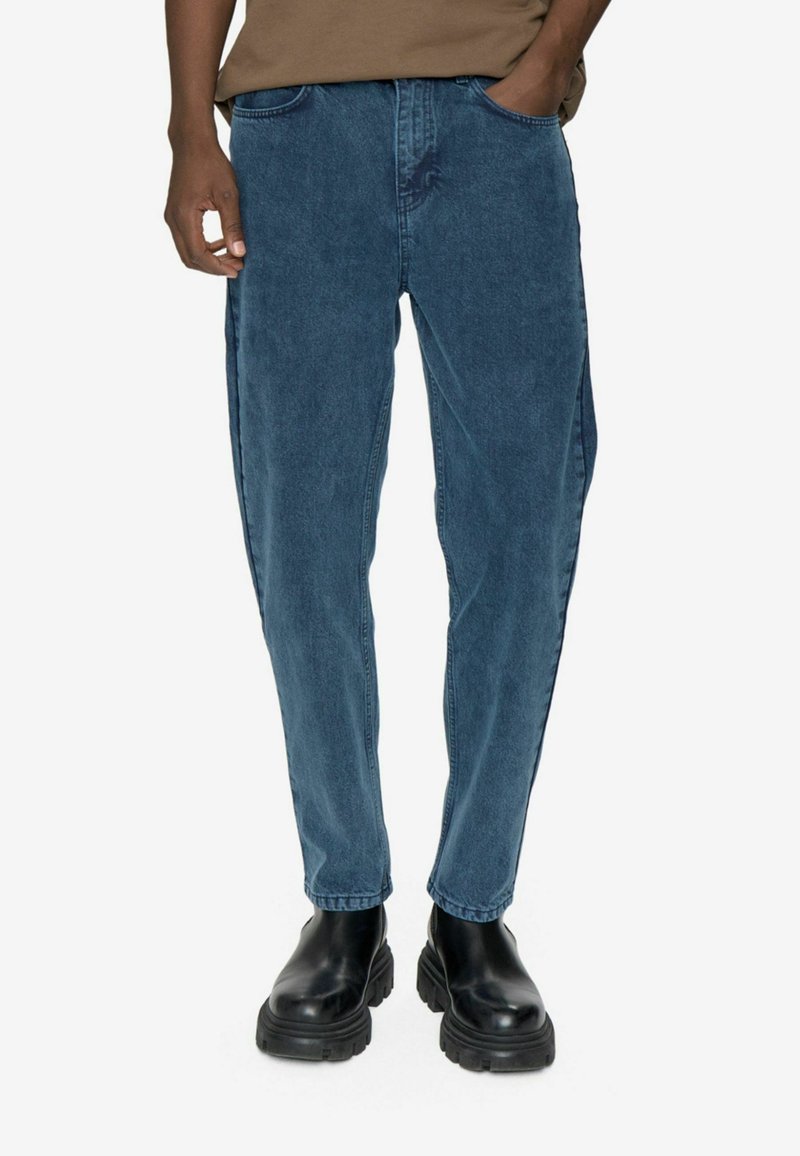 Modri denim džins s sproščenim krojem, ki ima pet žepov in subtilno zbledelost. V kombinaciji z črnimi masivnimi gležnjarji.