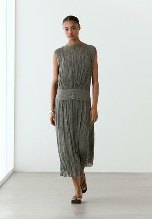 Donna che cammina con un vestito grigio testurizzato senza maniche con vita plissettata, sandali neri e braccialetto bianco su sfondo bianco semplice.