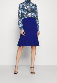 High-waisted blue pencil skirt with a flared bottom, paired with a sheer black blouse featuring a floral blue pattern and long sleeves.