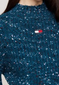 Close-up van een gestructureerde blauwe gebreide trui met multikleurige stippen en een rechthoekig Tommy Jeans-logo op de borst.