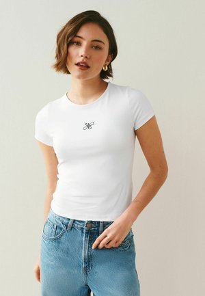 Jonge vrouw met kort bruin haar die een witte aansluitende T-shirt draagt met een klein zwart embleem en lichtblauwe spijkerbroek.