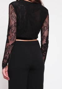 Top corto in pizzo nero con maniche lunghe trasparenti e schiena nera unita; abbinato a pantaloni neri a vita alta con cerniere laterali.