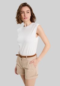 Weißes ärmelloses Oberteil mit rundem Ausschnitt, kombiniert mit beigen, taillierten Shorts mit Fältchen und einem geflochtenen braunen Gürtel.