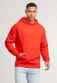 Felpa arancione realizzata in materiale morbido, con tasca frontale, cappuccio con cordino e polsini a coste. Indossata con jeans azzurri chiari.